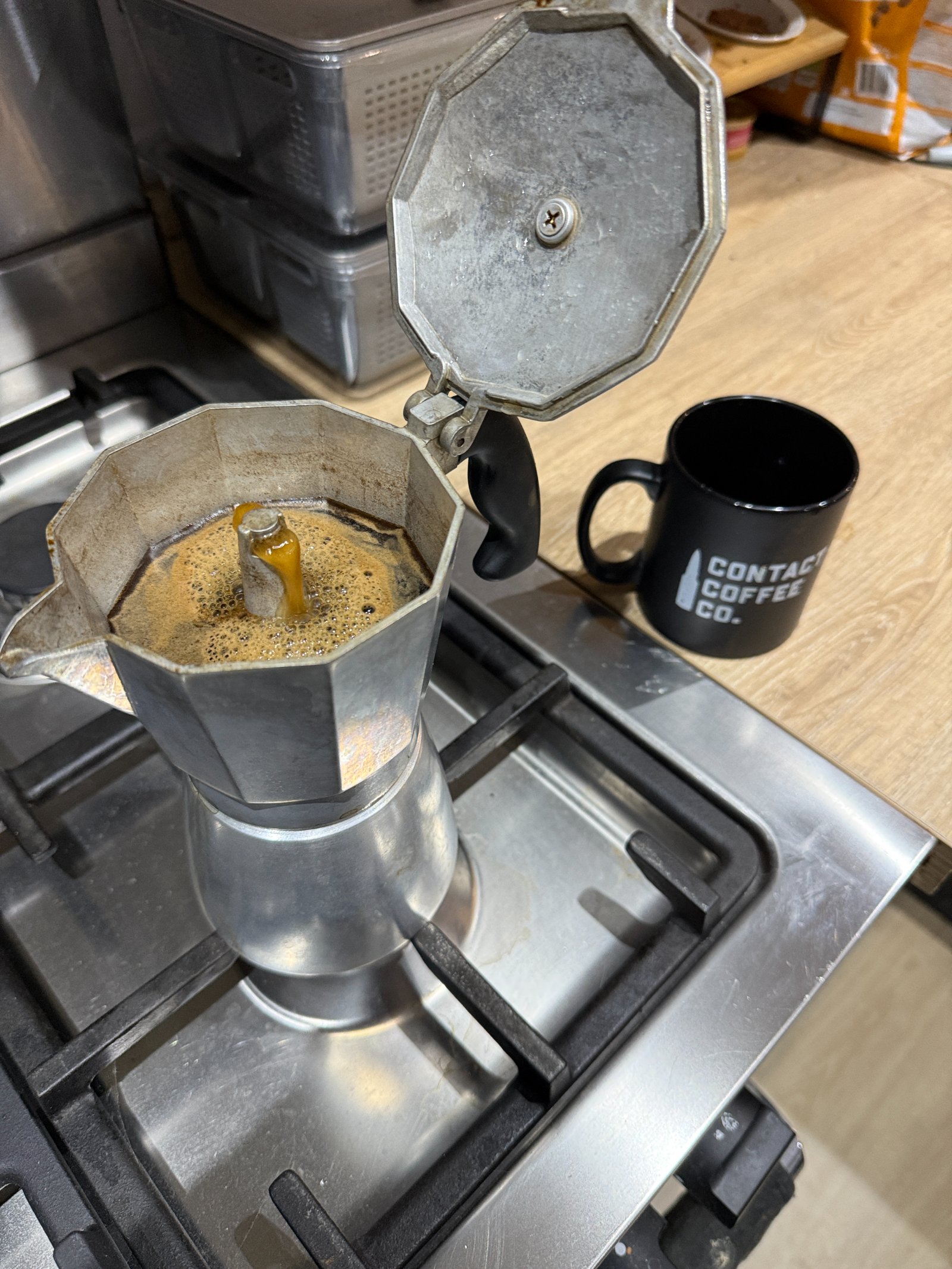 A moka pot heating on a kitchen hob in early morning light, with a plain mug and a bag of coffee on the worktop